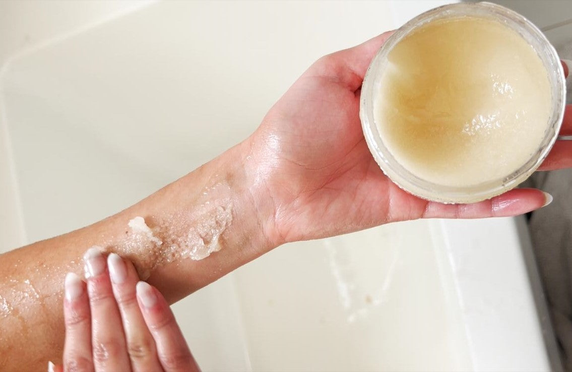 "Close-up of a hand applying a natural sugar body scrub to the wrist, revealing soft and smooth skin. A jar of exfoliating scrub is held in the other hand, showcasing its rich, hydrating texture."